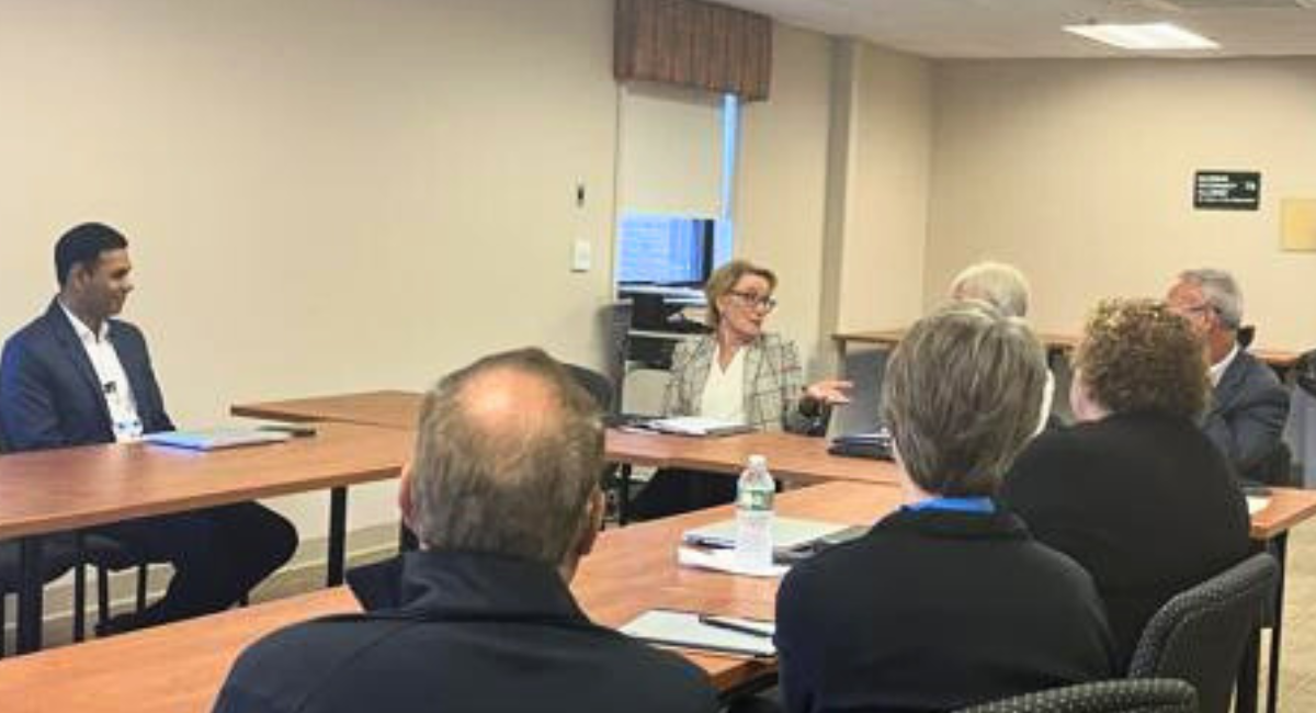 A group of healthcare and community leaders sit around conference tables in discussion, with one woman speaking while others listen during a meeting about maternal healthcare and delivery unit challenges.