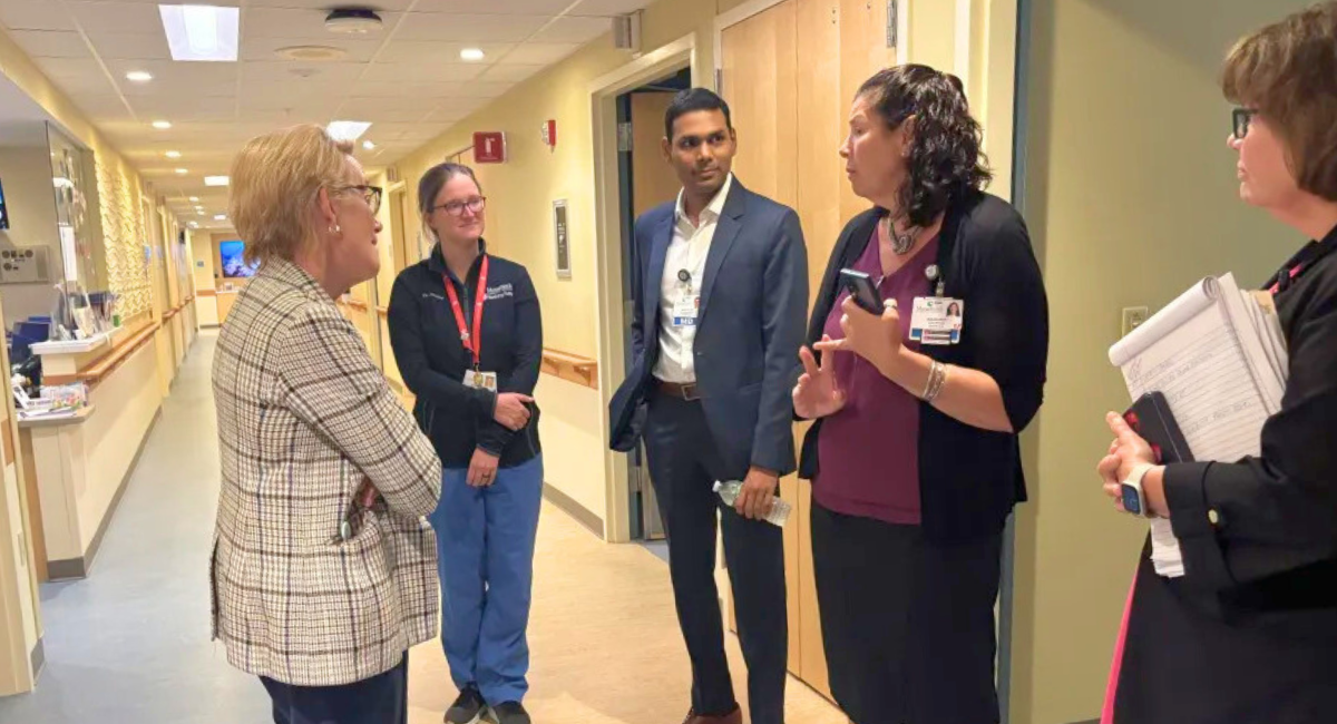 Group of five people standing in a hospital hallway having a discussion. A woman in a plaid jacket is facing four hospital staff members, including one in blue scrubs, one in a suit, and two in business attire with ID badges and notepads.