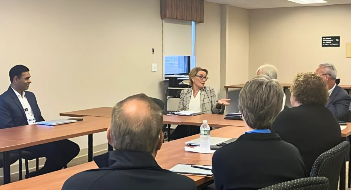 A group of healthcare and community leaders sit around conference tables in discussion, with one woman speaking while others listen during a meeting about maternal healthcare and delivery unit challenges.