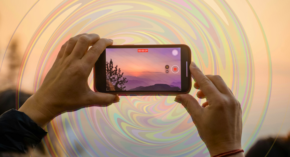 Hands holding a smartphone horizontally to record a sunset over mountains, with the phone screen showing a vivid orange and purple sky. A swirling rainbow pattern overlays the background