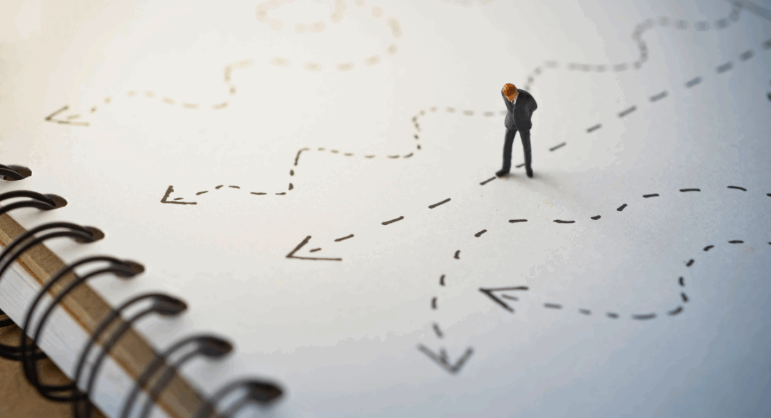A small figurine of a person in a suit stands on a notepad at a crossroads of dotted arrows, symbolizing choices and decision-making for the future.