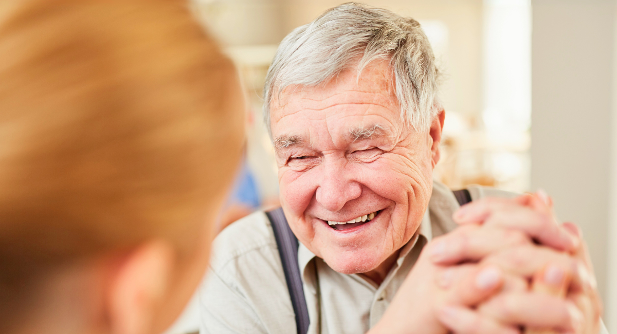 Close-up of an older man with gray hair smiling warmly, hands clasped, in conversation with a caregiver.