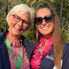 Two smiling women, one in a striped shirt and one in a navy MCH polo with a pink lei, stand together outdoors.