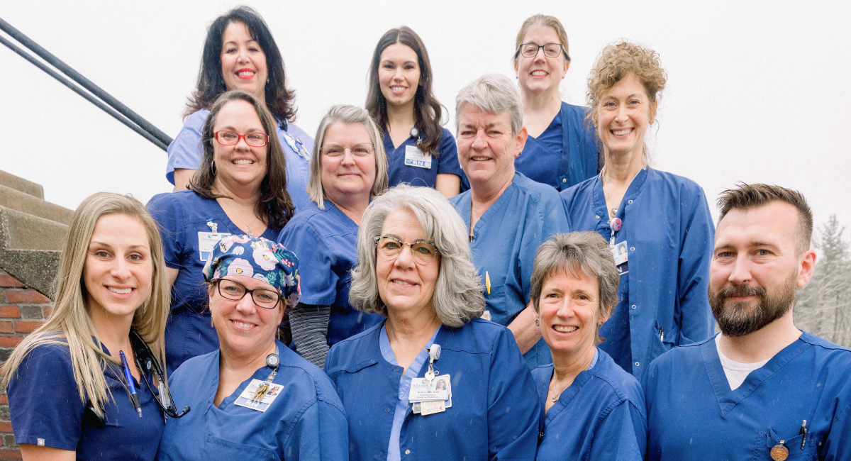 A group of Monadnock Community Hospital nurses stand together outdoors on a stairway, smiling at the camera. They are wearing blue scrubs and ID badges, showing a diverse team of men and women gathered in support of patient care.