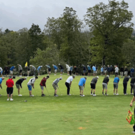 Dozens of golfers lined up in a large circle on the green, bending over their putters in a fun, synchronized moment.