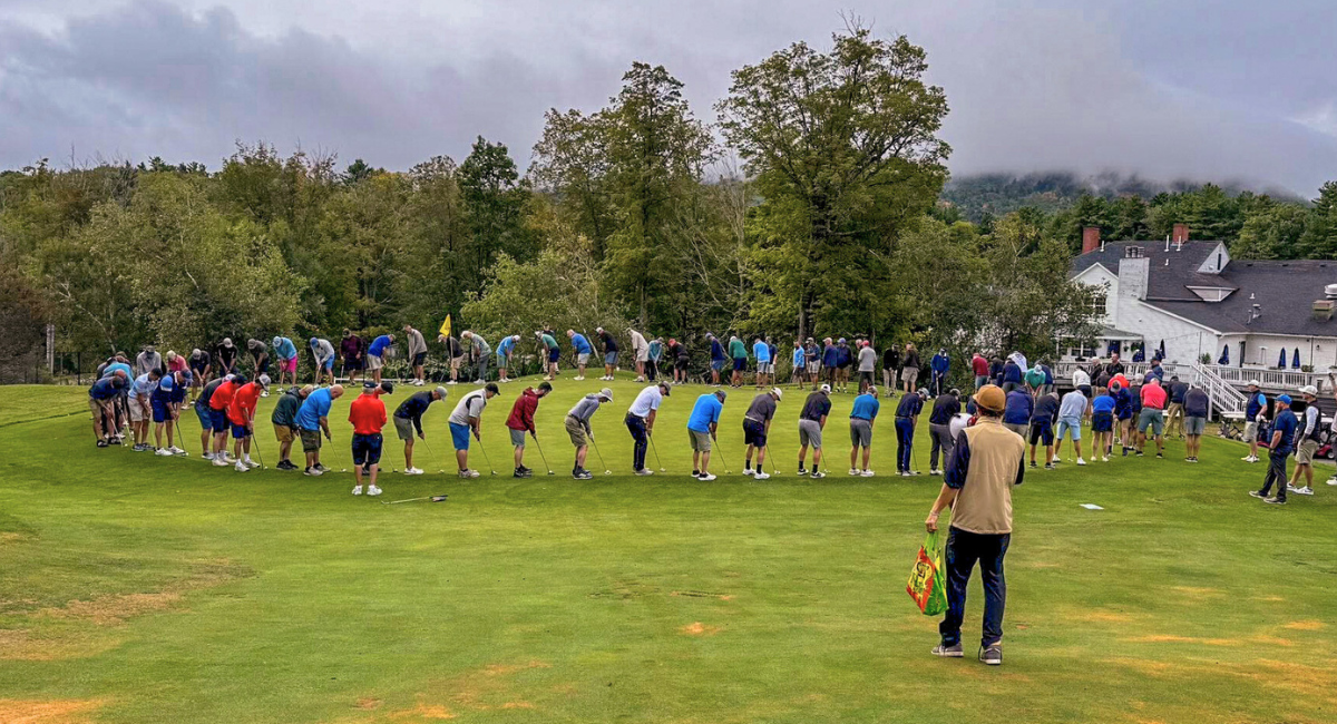 Large group of golfers standing in a wide circle on a green, all bent over preparing to putt, with a clubhouse and trees in the background under cloudy skies.