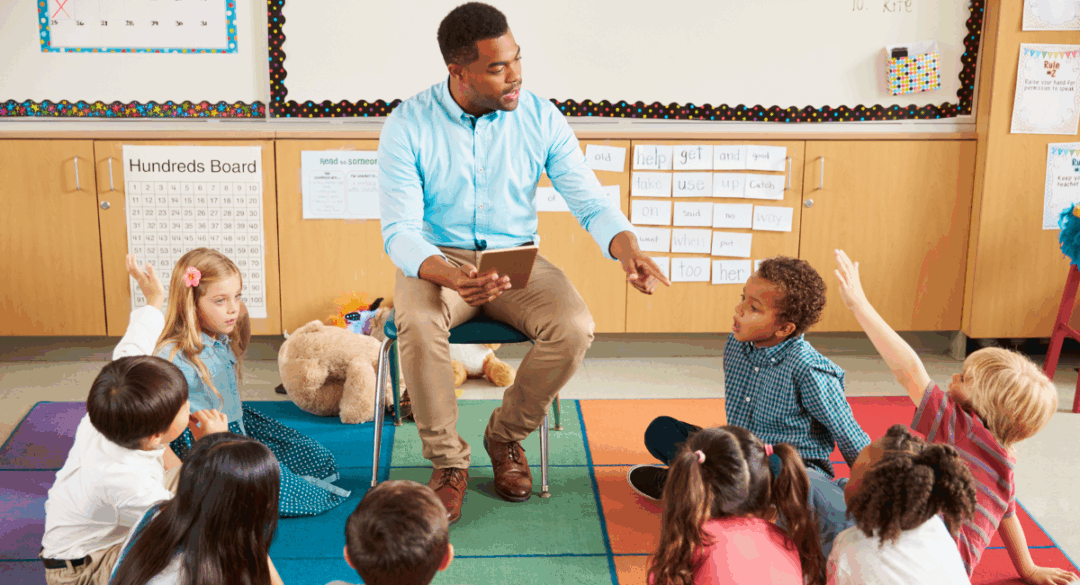 Teacher sitting on a chair holding a tablet, surrounded by young students sitting on a colorful classroom rug, with some children raising their hands to answer a question.