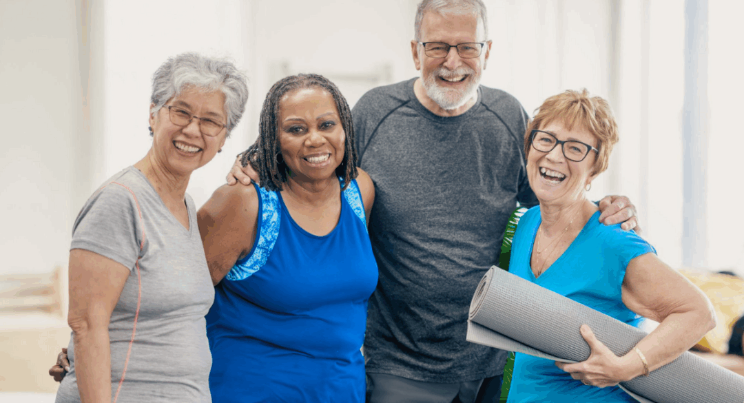 A group of four senior adults, two women and two men, smiling and standing close together in a bright, indoor setting. One woman is holding a yoga mat, indicating they are possibly participating in a wellness or fitness activity.