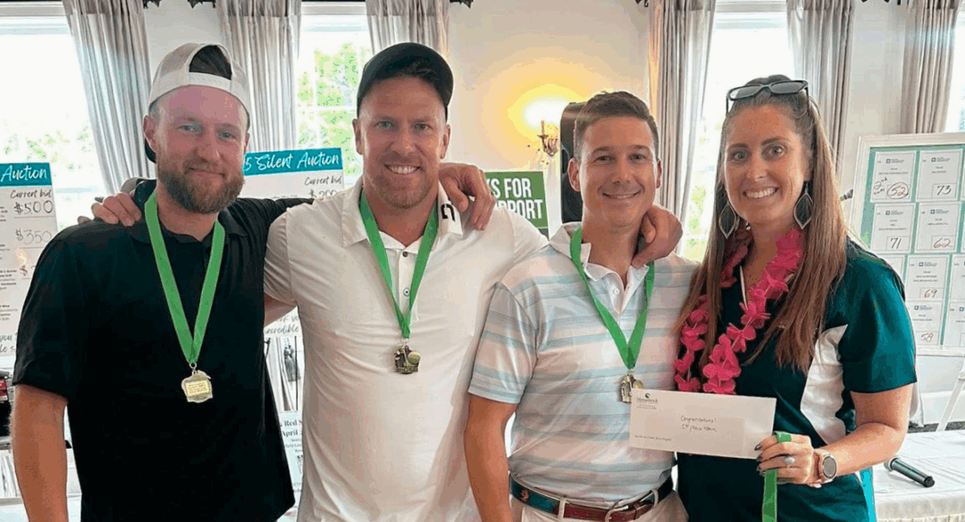Four golfers, wearing medals and holding their prize envelope, smile together after winning the tournament.