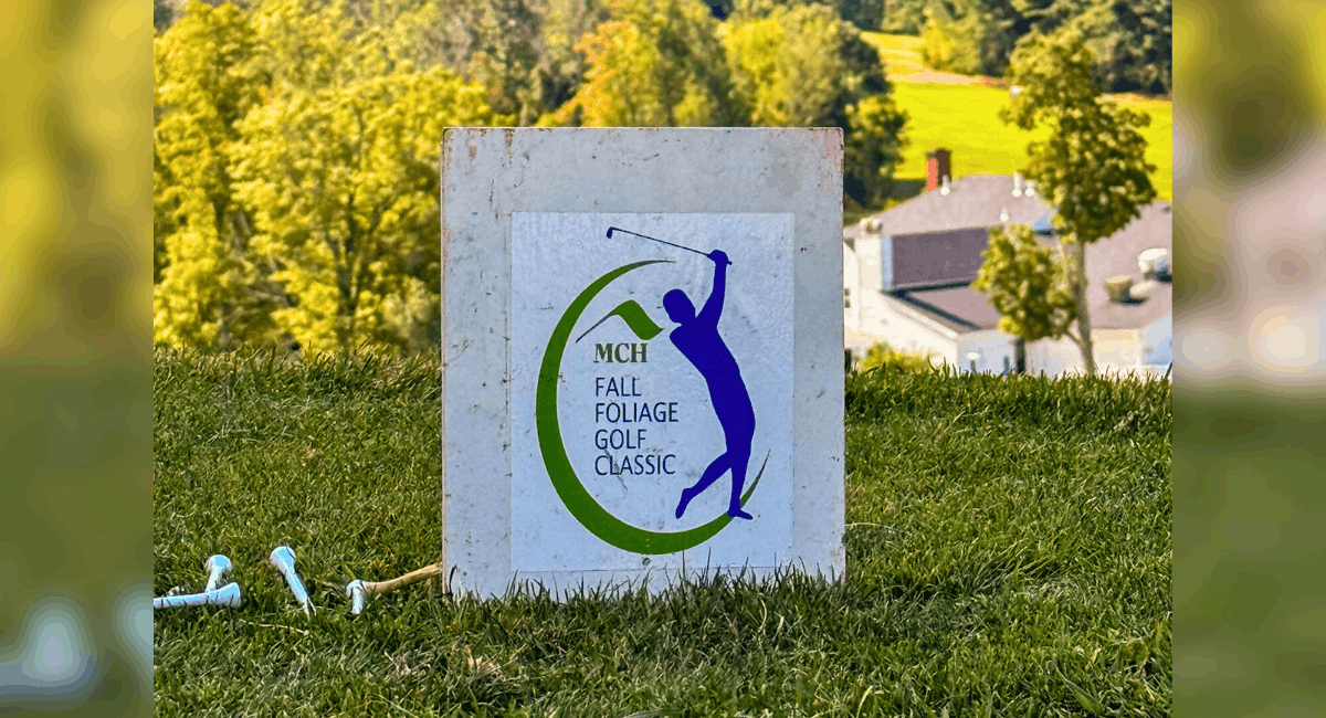 Close-up of a tee marker sign with the logo for Monadnock Community Hospital’s Fall Foliage Golf Classic, set against a scenic backdrop of rolling green hills and forest.