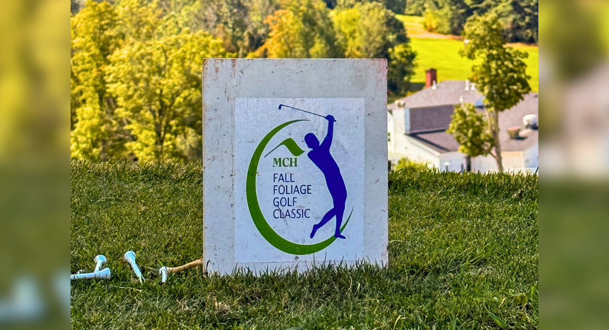 Close-up of a tee marker sign with the logo for Monadnock Community Hospital’s Fall Foliage Golf Classic, set against a scenic backdrop of rolling green hills and forest.