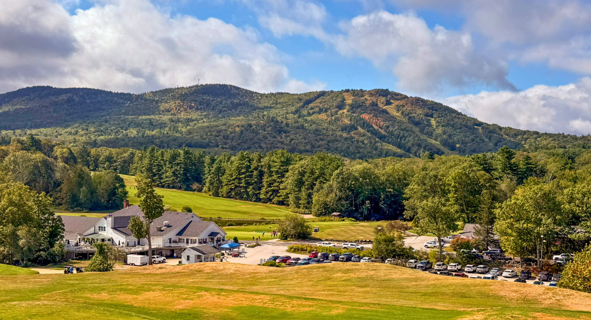 Panoramic view of a golf course and clubhouse surrounded by lush trees, with Mount Monadnock rising in the background on a partly cloudy day.