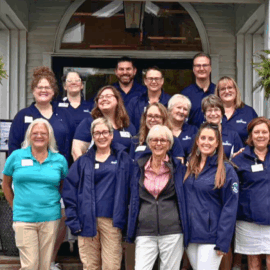 A large group of Monadnock Community Hospital volunteers and staff pose together outside the clubhouse, smiling in matching jackets.