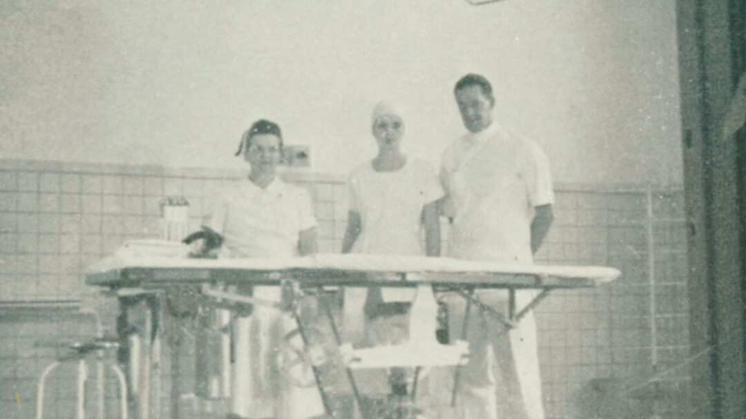 Three Monadnock Community Hospital surgical staff members standing beside an operating table in a 1940s operating room, wearing white uniforms and caps, representing the hospital’s long tradition of skilled, compassionate care.