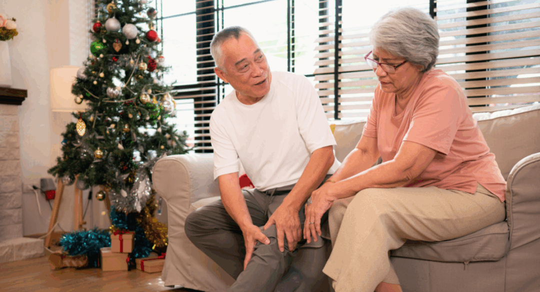 Older adult couple sitting on a couch near a Christmas tree; the woman rubs her knee in discomfort while the man offers support, symbolizing joint pain during the holidays.
