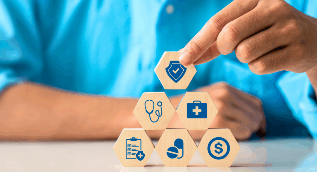 A person in a blue shirt stacks wooden hexagon blocks on a table, each printed with a blue healthcare icon including a stethoscope, medical bag, checklist, pill, dollar sign, and shield symbol, representing health coverage and insurance protection.
