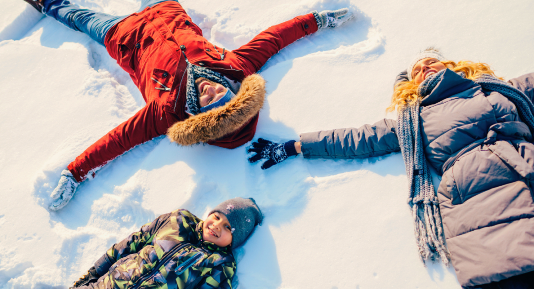 Three people in winter coats lie in fresh snow making snow angels, capturing a joyful, relaxed moment of winter outdoor activity and seasonal wellness.