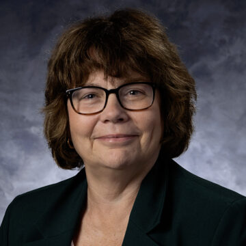 Professional headshot of a woman wearing glasses and a dark green blazer against a gray studio background