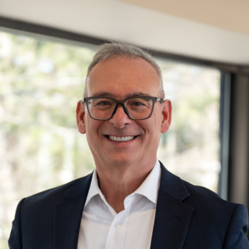 Richard Scheinblum smiling in a suit and glasses in front of a window with a softly blurred background