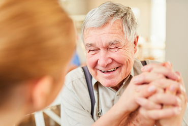 lose-up of an older man with gray hair smiling warmly, hands clasped, in conversation with a caregiver.
