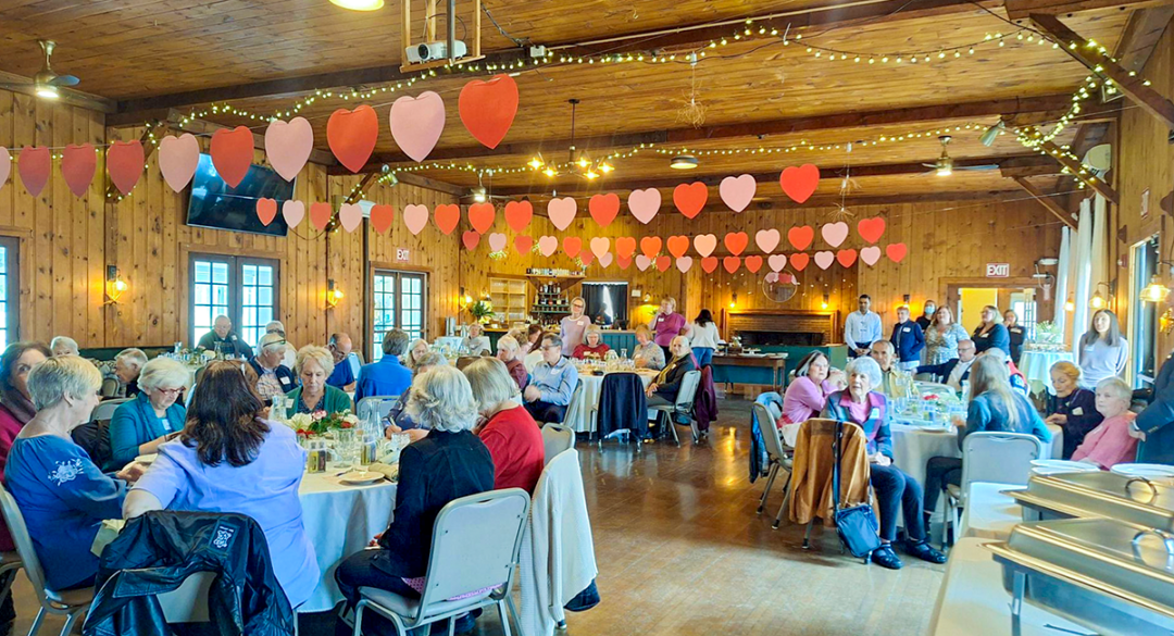 Monadnock Community Hospital volunteers gather for the annual Volunteer Luncheon in a warmly decorated banquet hall. Guests sit at round tables enjoying lunch and conversation beneath strings of red and pink heart decorations hanging across the wooden-beamed ceiling.