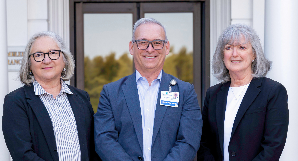 President & CEO Richard Scheinblum stands outside Monadnock Community Hospital with Board Chair Paula Hunter on the left and Vice Chair Linda Roche on the right. All three are dressed in business attire and smiling in front of the hospital entrance.