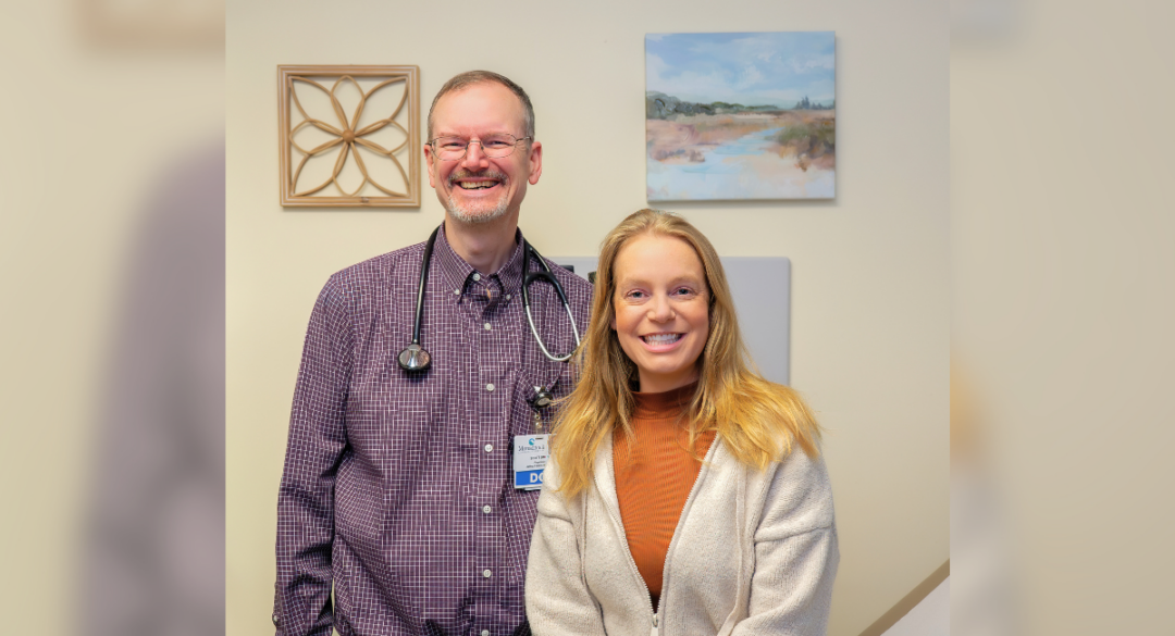 Scott E. Smith, DO, stands beside Christine TenBroeck in an exam room at Jaffrey Family Medicine. Christine sits on the exam table and smiles as they pose together for a portrait.