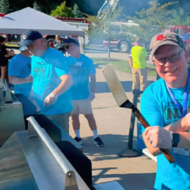 National Night Out volunteers prepare food at an outdoor grill with fire trucks and a raised American flag in the background.
