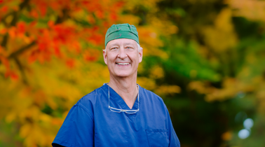 Smiling older man in blue scrubs and a green scrub cap with reading glasses resting on his chest, standing in front of red and yellow foliage.